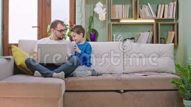 年轻的爸爸和他的小儿子一起在沙发上度过快乐时光，他们在笔记本电脑上玩，享受着时光